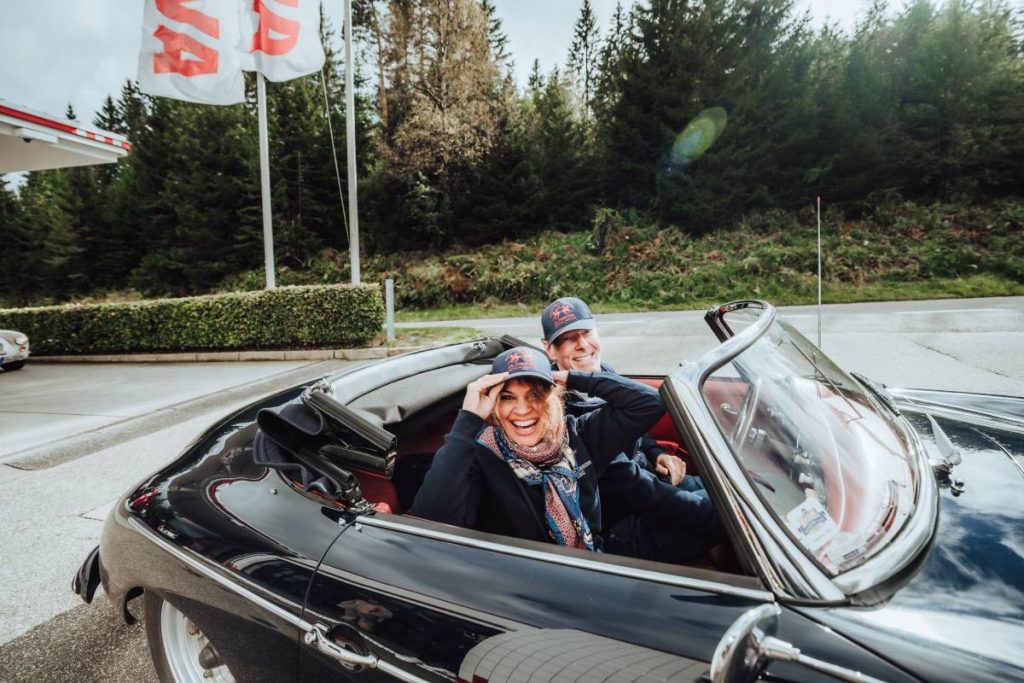 Events - a good mood in Baiersbronn: a good-looking middle-aged couple sits in a black vintage convertible with the top down and laughs at the camera. Both are wearing the same blue cap.