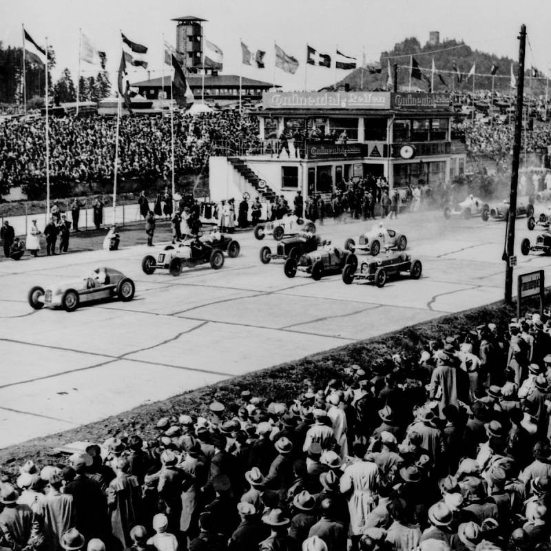 Legendäres Event: Heißer Kampf beim Eifelrennen 1934: Auf dem Bild sind viele Oldtimer-Cabrios zu sehen, die um den Sieg beim Eifelrennen kämpfen - vorneweg der Silberpfeil von Mercedes-Benz. Viele Zuschauer stehen gespannt auf den Tribünen.