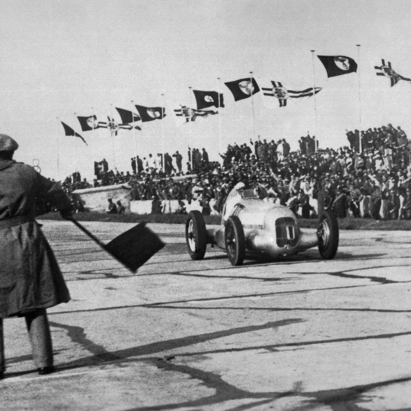 Schwarz-weiß-Bild: Manfred von Brauchitsch gewinnt 1934 im Silberpfeil von Mercedes-Benz das Eifelrennen. Im Vordergrund steht der Mann mit der Zielflagge. Im Hintergrund sieht man die Zuschauertribüne.