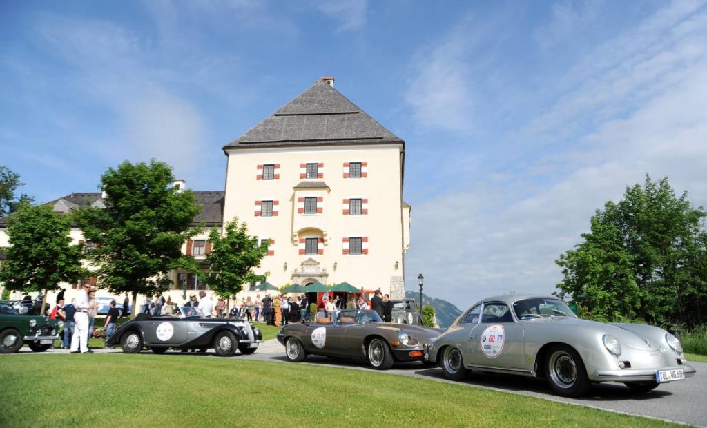 Lifestyle meets Drivestyle: Vor dem Schloss Fuschl stehen wertvolle Oldtimer, die an der ArabellaClassics Rallye teilnehmen. Die Türen der Fahrzeuge sind mit Startnummern beklebt. Menschen drängen sich vor dem Schloss und um die Fahrzeuge.