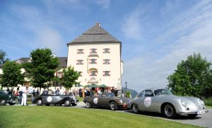 Vor dem Schloss stehen wertvolle Oldtimer, die an der ArabellaClassics Rallye teilnehmen. Die Türen der Fahrzeuge sind mit Startnummern beklebt. Menschen drängen sich vor dem Schloss und um die Fahrzeuge.