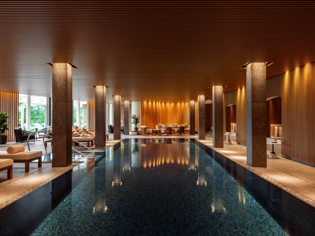 Schloss Fuschl steht für Lifestyle: Man sieht den Spa-Bereich mit edlem Interieur in Beige- und Brauntönen. Den Mittelpunkt bietet der geradlinige Pool, der von eckigen Säulen umrahmt wird. Auf dem Wasser spiegeln sich Lichter wider.
