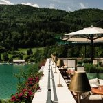 Auf der Seeterrasse von Schloss Fuschl stehen bequeme Stühle und Sessel sowie beige Sonnenschirme. Von dort blickt man direkt auf den smaragdgrünen Fuschlsee und das Ufer mit Booten. Die Terrasse ist mit edlen Accessoires geschmückt.