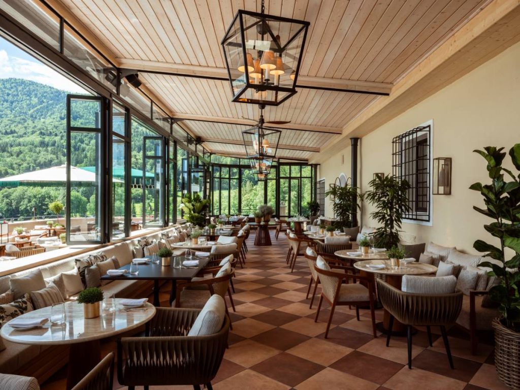Auf der Seeterrasse von Schloss Fuschl mit dem großen Wintergarten im Vordergrund stehen viele bequeme braune Stühle und Sessel mit weißen Kissen. Die Fenster zum See sind weit geöffnet. Im Hintergrund sieht man üppig grüne Vegeation.