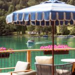 Auf der Seeterrasse stehen bequeme Sessel und ein blauer Sonnenschirm. Man blickt direkt auf den See und das gegenüberliegende Ufer mit Booten. Ein Boot mit Gästen nähert sich der mit Geranien-Kästen geschmückten Terrasse.