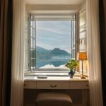 Durch ein geöffnetes Fenster blickt man vom Schloss auf den Fuschlsee und die dahinter aufragenden Berge. Auf dem kleinen Schreibtisch davor liegen zwei dünnen Bücher. Rechts auf dem Schreibtisch stehen eine Vase mit bunten Blumen und eine kleine Tischlampe.