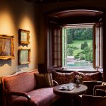 Der Salon ist durch warme Braun-, Rot- und Goldtöne geprägt. An der Wand hängen drei Gemälde mit goldenen Rahmen. Ein Fenster mit Rundbogen ist weit geöffnet und gibt den Blick frei auf die Bootsanlegestelle.