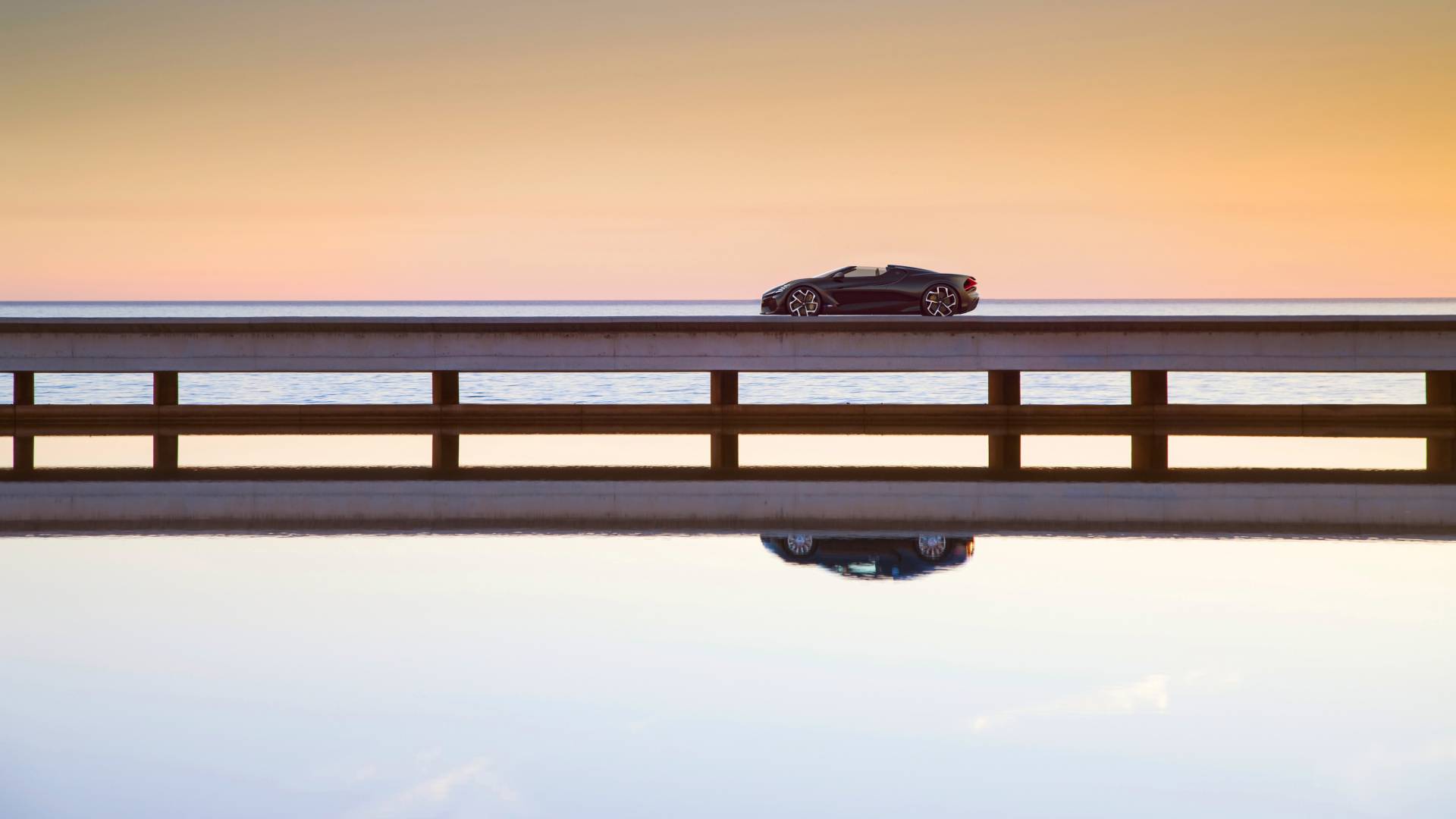 Ein schwarzer Bugatti W16 Mistral fährt auf einer langgestreckten Brücke von rechts nach links. Darunter sieht man eine glatte Wasseroberfläche auf der sich das Cabrio widerspiegelt. Das Bild ist in warmen Beige- und Brauntönen gehalten.