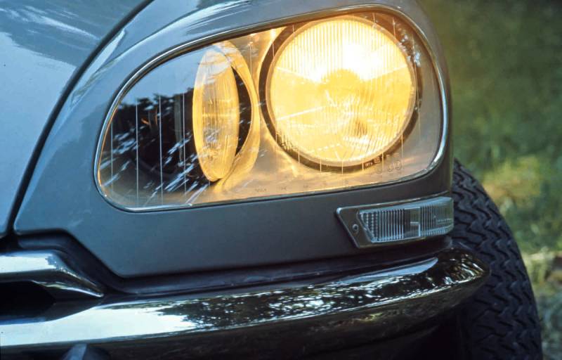 Ein Teil der Front einer blauen Citroen DS mit gelbem Scheinwerfer ist zu sehen. Das rechte Licht ist voll nach vorne fokussiert. Das linke Licht ist zur Hälfte zu sehen. Am unteren Rand sieht man die silbern glänzenden Stoßstange.