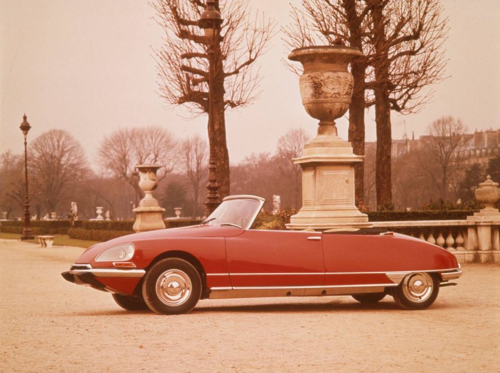 Eine rotes Cabrio der Marke Citroen DS steht mit geöffnetem Verdeck auf einem Parkgelände in Paris. Die seitliche Aufnahme unterstreicht die eindrucksvolle Formgebung des Cabrios. Das Foto stammt aus den 60er-Jahren und ist schon etwas verblichen.