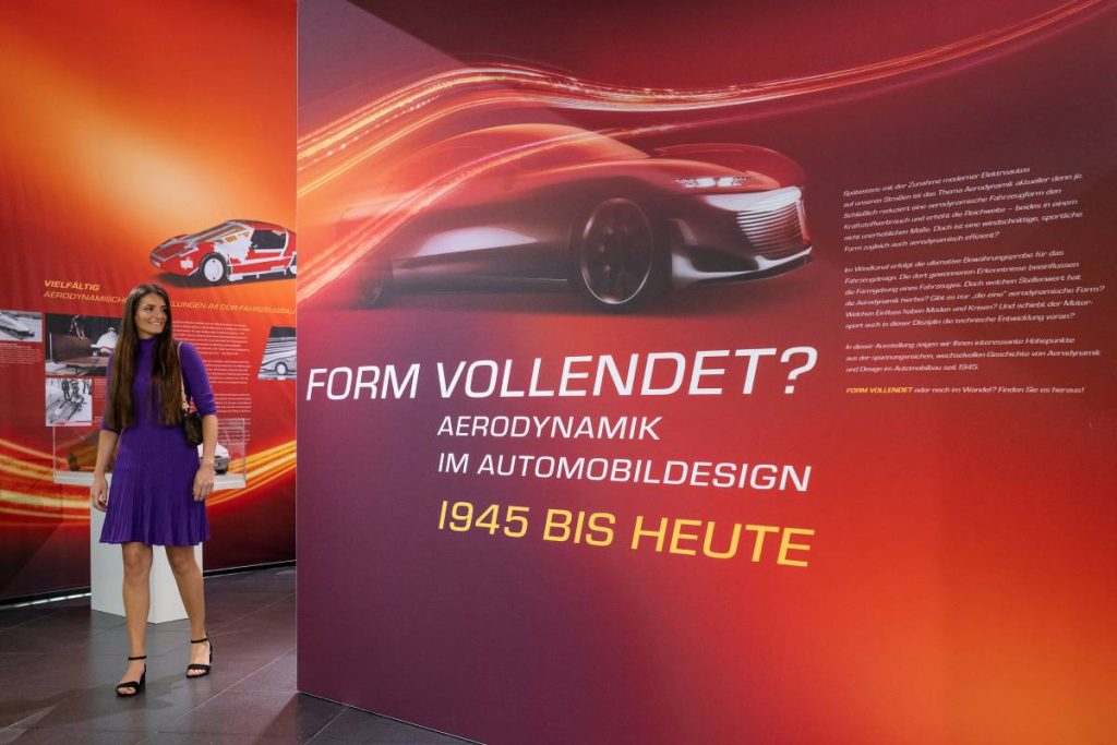 Event for convertible drivers: A poster in the colors orange to dark red and a young visitor in a blue dress. The poster from Audi Tradition advertises the new special exhibition "Form completed" at the Audi museum mobile.