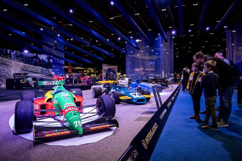 Es wird die Ausstellungshalle der InterClassics Maastricht mit blauem Teppichboden mit vielen historischen offenen Sportwagen gezeigt. Ein Vater mit seinen zwei Kindern betrachtet einen grünen Rennwagen mit der Nummer 20.