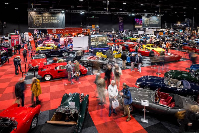 Es wird eine Halle der InterClassic Maastricht mit rotem Teppichboden gezeigt. Im Vordergrund stehen glänzende offene Oldtimer in allen möglichen Farben, die von zahlreichen Zuschauern betrachtet werden.