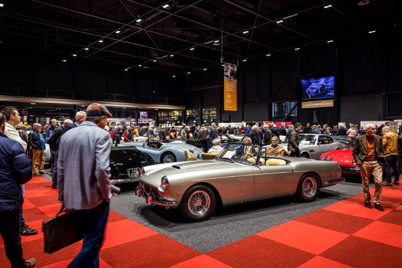 Es wird eine Halle der InterClassic Maastricht mit rotem Teppichboden gezeigt. Im Mittelpunkt steht ein silberner glänzender offener Oldtimer, der von allen Seiten von Zuschauern betrachtet wird.