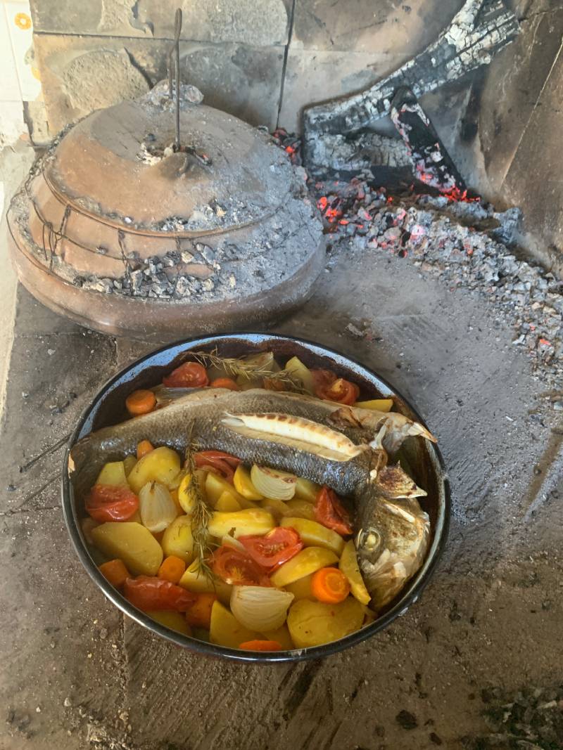Es sind eine Tonglocke und eine große Pfanne mit gebratenem Fisch und Gemüse sowie dahinter Reste von Glut und Asche zu sehen. In Kroatien werden Speisen in der sogenannten "Peka" (Glocke) zubereitet.