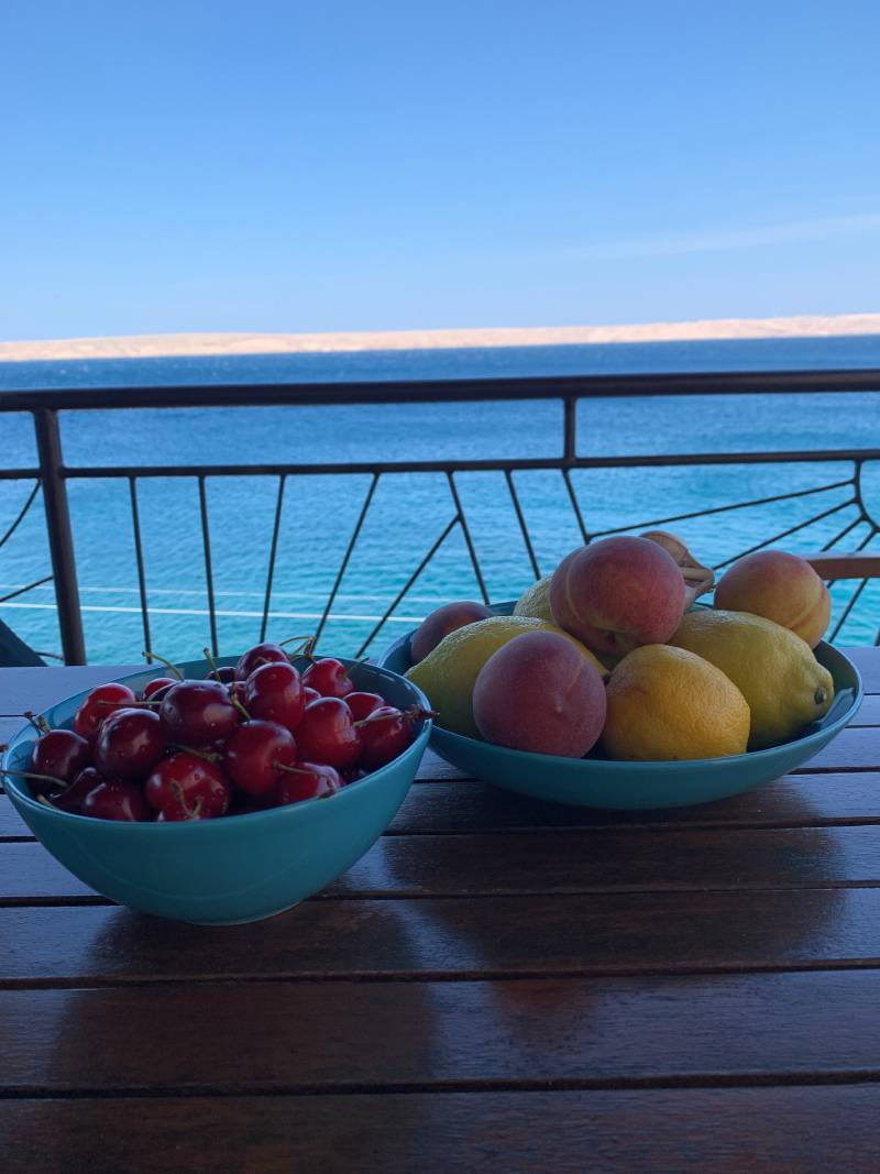 Cabriolife on Tour: Auf dem Tisch auf einem Balkon stehen zwei Schüsseln mit Kirschen und anderem frischen Obst. Im Hintergrund sieht man das azurblaue Meer und einen weißen Streifen – die Insel Pag.