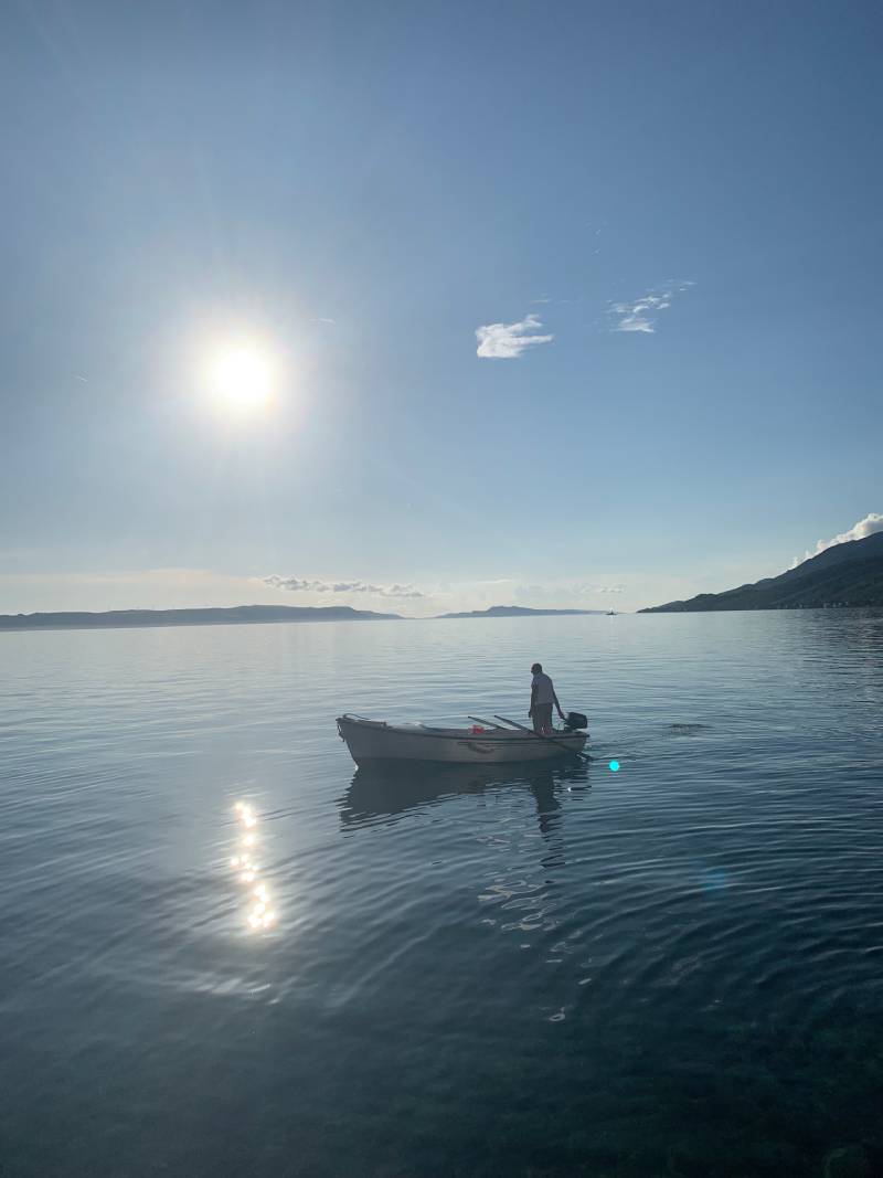 Auf dem Wasser fährt ein Fischer mit seinem kleinen Boot im Sonnenuntergang vor der Küste. Die runde Sonne leuchtet silbern vom Himmel herab.