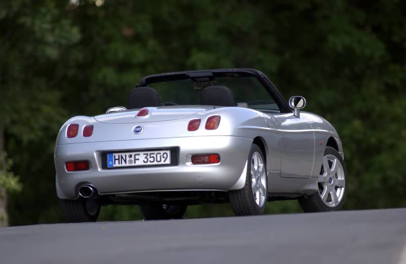 A silver Fiat Barchetta convertible is seen diagonally from behind with the roof open.