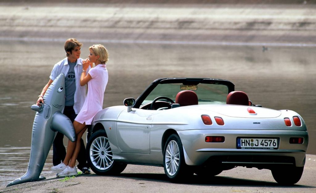 A young couple on the beach lean against a silver Fiat Barchetta and cuddle.