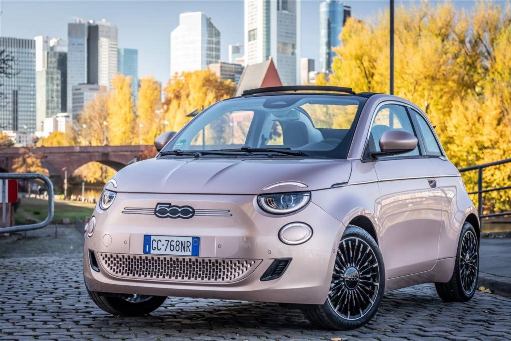 Ein FIAT 500e Cabrio in der Farbe Rosé-Gold steht auf einer Straße. Im Hintergrund sieht man herbstlich gefärbte Bäume und die Skyline einer modernen Stadt.
