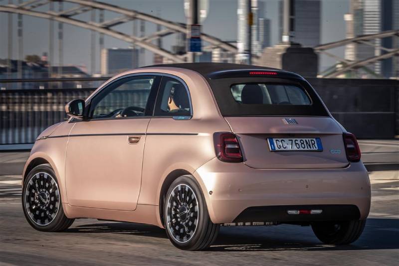 Ein FIAT 500e Cabrio in der Farbe Rose-Gold fährt durch eine Großstadt. Im Hintergrund sieht man eine große Brücke und die Skyline einer Stadt am Tag.