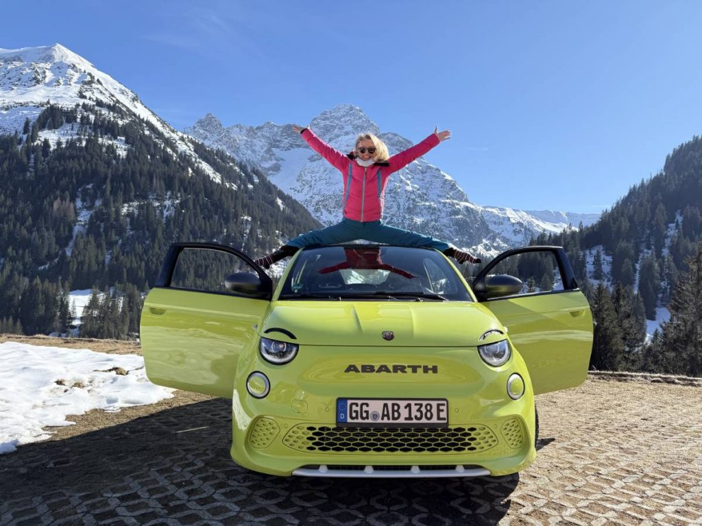 Abarth 500e in der Farbe Neongrün mit einer jungen Frau, die Spagat auf dem Cabrio macht. Im Hintergrund sieht man schneebedeckte Berge und stahlblauen Himmel.