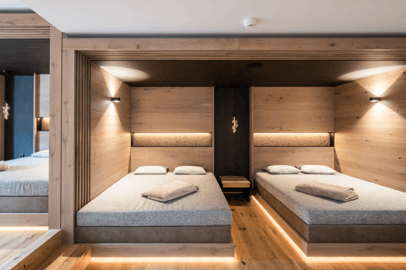 Man sieht zwei einzelne Betten nebeneinander. Die Wände des Schlafzimmers sind aus warmem Holz. Auf der linken Seite gibt es eine Spiegelung des Raums. Das Bild ist in Braun- und Grautönen gehalten und wird von Lichtstrahlen komplettiert.