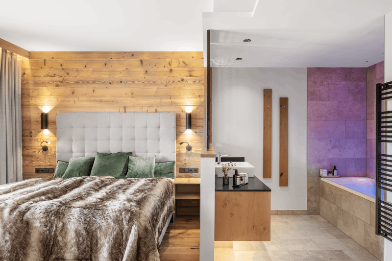 Auf der linken Seite sieht man ein großes Bett mit Felldecke und rustikaler Holzwand als Hintergrund. Auf der rechten Seite sieht man das angrenzende moderne Bad mit Holz und Naturstein. Das Bild ist in warmen Brauntönen gehalten.