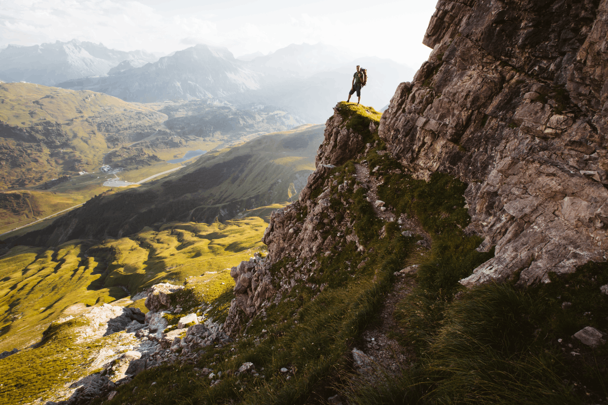 Ein Bergsteiger blickt von einem exponierten Felsvorsprung am Großen Widderstein hinunter ins Tal. Im grünen Tal sieht man einen Fluss und zwei blaue Bergseen. Im Hintergrund sind die Lechtaler Alpen.