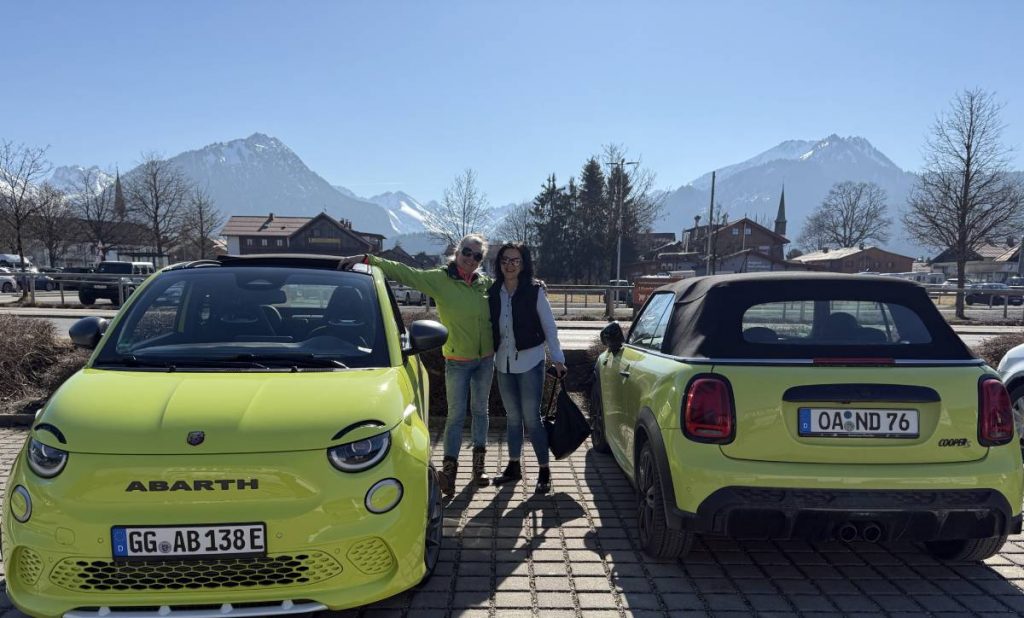 Zwei junge Frauen stehen zwischen zwei neongrünen Cabrios. Links ist der Abarth 500e von vorne und rechts ein BMW Mini Cabrio von hinten zu sehen. Im Hintergrund sieht man schneebedeckte Berge und stahlblauen Himmel.