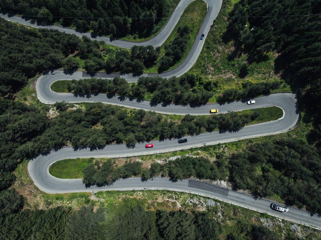 Man sieht eine Luftaufnahme von einer bewaldeten Bergstraße, auf der bunte Autos fahren. Die Straße führt über vier Kehren durch eine grüne Landschaft. Grün- und Blautöne beherrschen das Bild.