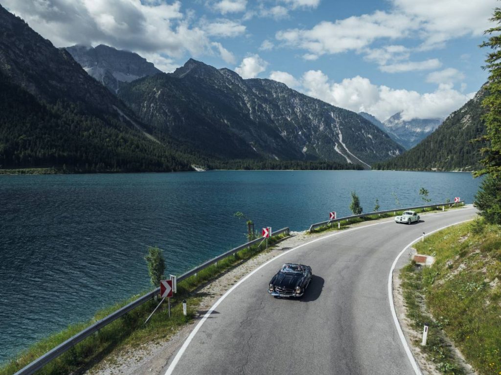 Ein schwarzes Mercedes-Benz Oldtimer-Cabrio fährt bei der "16. Arlberg Classic Car Rally" auf einer Straße am See entlang. Dahinter fährt ein anderer weißer Oldtimer. Im Hintergrund sieht man schroffe Berge. Grün- und Blautöne beherrschen das Bild.