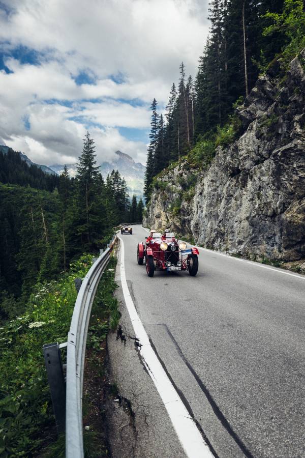 Zwei sehr alte Oldtimer-Cabrio fahren bei der "16. Arlberg Classic Car Rally" hintereinander auf einer Bergstraße. Auf der rechten Seite sieht man Felswände. Grau- und Grüntöne beherrschen das Bild.