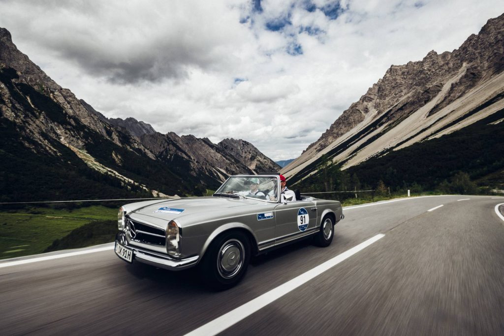 Ein silbernes Mercedes-Benz Oldtimer-Cabrio fährt bei der "16. Arlberg Classic Car Rally" auf einer Bergstraße. Im Hintergrund sieht man schroffe Berge und viele Wolken. Grau- und Brauntöne beherrschen das Bild.