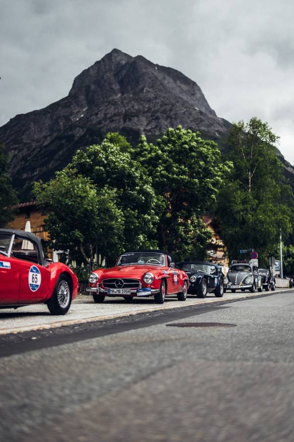 Viele Oldtimer-Cabrios stehen am Straßenrand eines Ortes bei der "16. Arlberg Classic Car Rally". Im Hintergrund sieht man einen hohen Berg und grüne Bäume. Grau- und Rottöne beherrschen das Bild.
