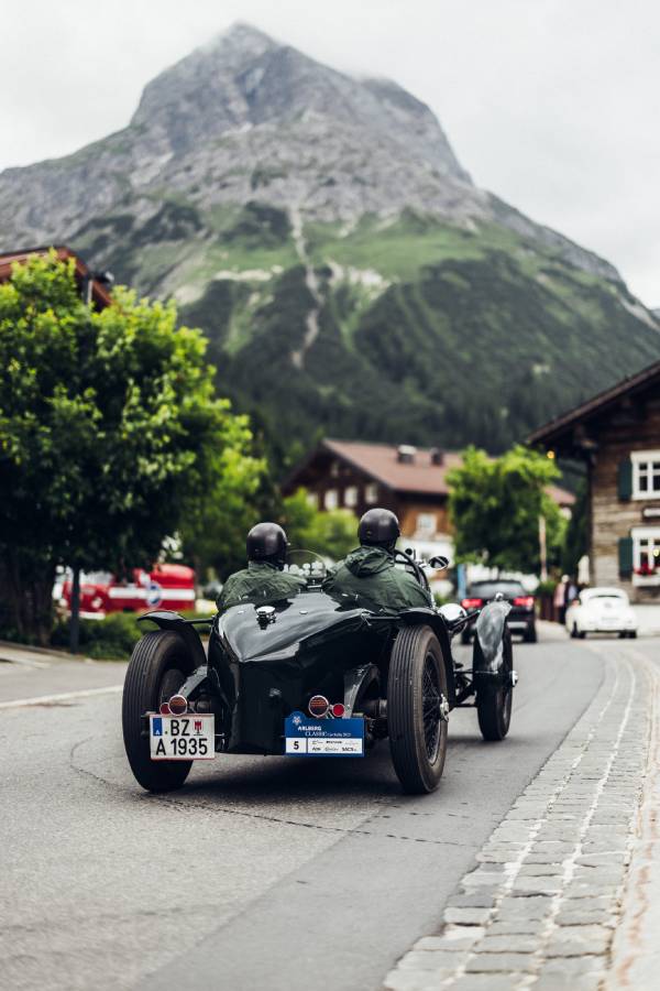 Ein schwarzes Oldtimer-Cabrio fährt bei der "16. Arlberg Classic Car Rally" auf einer Straße in einen Ort hinein. Im Hintergrund sieht man einen hohen Berg und grüne Bäume. Grau- und Grüntöne beherrschen das Bild.
