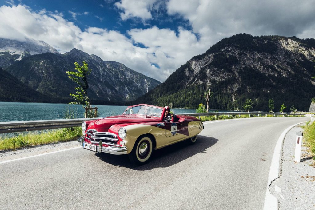 Ein zweifarbiges Oldtimer-Cabrio (rot-gelb) fährt bei der "16. Arlberg Classic Car Rally" auf einer Straße am See entlang. Im Hintergrund sieht man schroffe Berge. Grau- und Grüntöne beherrschen das Bild.