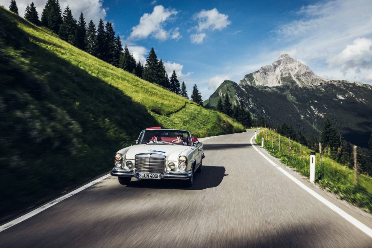 Ein weißes Oldtimer-Cabrio fährt bei der "16. Arlberg Classic Car Rally" auf einer Bergstraße. Im Hintergrund sieht man felsige Berge und grüne Hänge. Grau- und Grüntöne beherrschen das Bild.