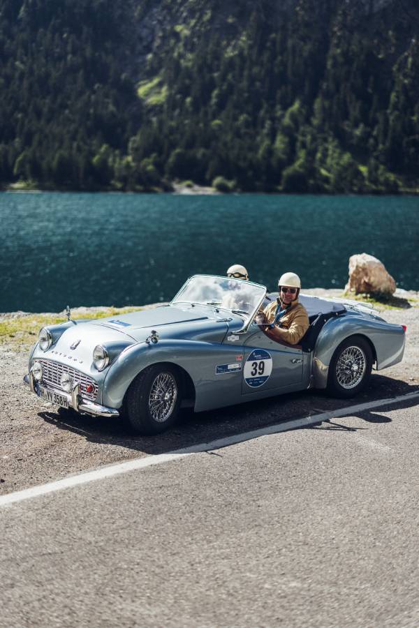 Ein silbernes Oldtimer-Cabrio fährt bei der "16. Arlberg Classic Car Rally" auf einer Straße am See entlang. Fahrer und Beifahrer tragen Schutzhelme. Im Hintergrund sieht man grüne Berge. Blau- und Grüntöne beherrschen das Bild.