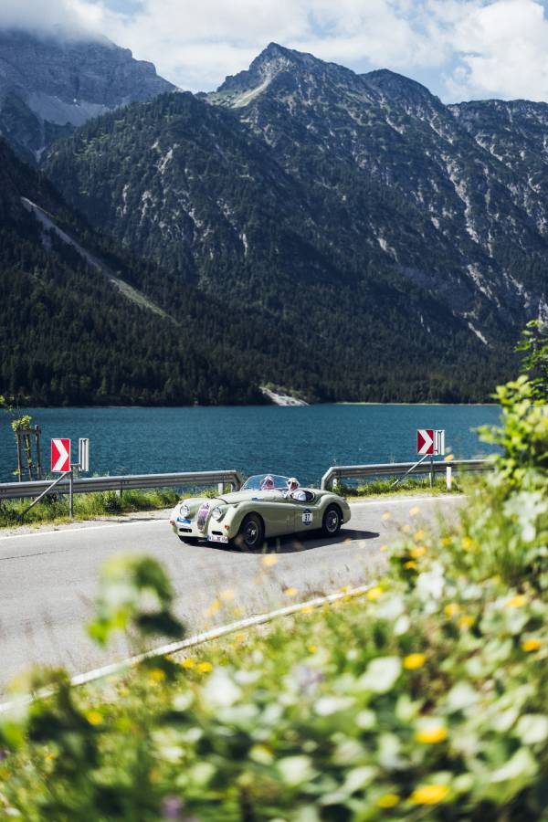 Ein silbernes Oldtimer-Cabrio fährt bei der "16. Arlberg Classic Car Rally" auf einer Straße am See entlang. Im Hintergrund sieht man hohe Berge. Blau- und Grüntöne beherrschen das Bild.