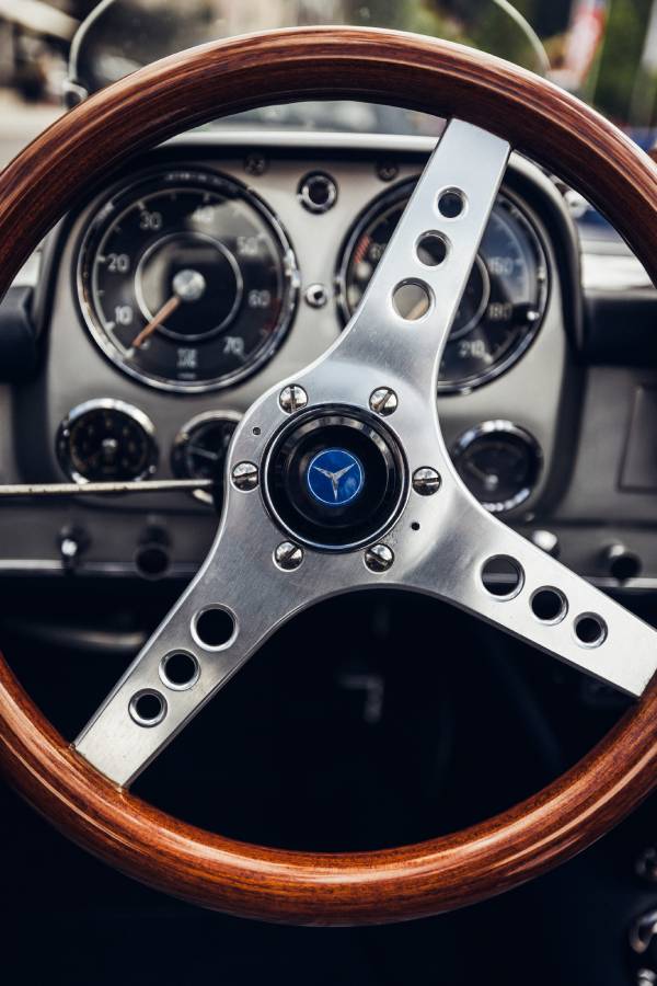Man sieht die Nahaufnahme eines Lenkrad aus Holz mit drei gelöcherten Metallspeichen und dahinter das Armaturenbrett. In der Mitte des Lenkrads prangt ein Stern – das Emblem von Mercedes-Benz.