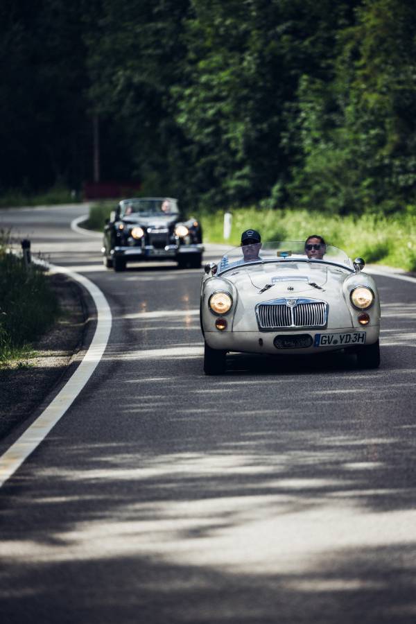 Zwei Oldtimer-Cabrio fahren bei der "16. Arlberg Classic Car Rally" hintereinander durch einen Nadelwald auf einer Bergstraße. Im Hintergrund sieht man einen hohen Berg. Grau- und Grüntöne beherrschen das Bild.