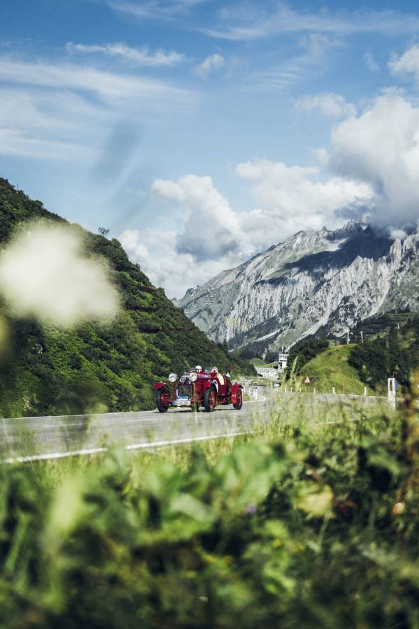 Ein rotes Oldtimer-Cabrio fährt bei der "16. Arlberg Classic Car Rally" auf einer Bergstraße. Im Hintergrund sieht man felsige Berge und grüne Hänge. Blau- und Grüntöne beherrschen das Bild.