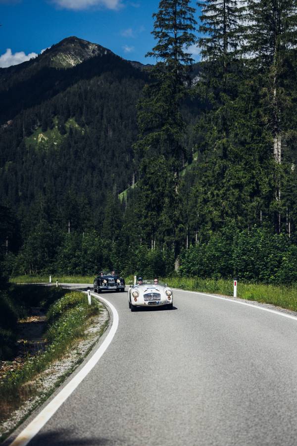 Zwei Oldtimer-Cabrio fahren bei der "16. Arlberg Classic Car Rally" hintereinander durch einen Nadelwald auf einer Bergstraße. Im Hintergrund sieht man einen hohen Berg. Grau- und Grüntöne beherrschen das Bild.