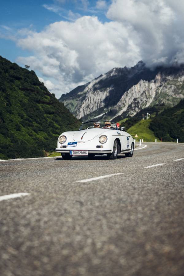 Ein weißes Oldtimer-Cabrio fährt bei der "16. Arlberg Classic Car Rally" auf einer Bergstraße. Im Hintergrund sieht man felsige Berge und grüne Hänge. Grau- und Grüntöne beherrschen das Bild.