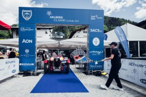 Ein sehr altes Oldtimer-Cabrio steht am Start der "16. Arlberg Classic Car Rally" auf blauem Teppich. Ein Mann gibt mit dem Schwenken einer Fahne das Zeichen zum Start. Blautöne beherrschen das Bild.