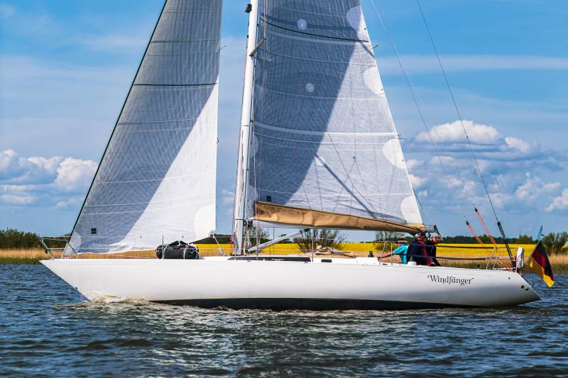 Omega 42 vom schwedischen Yacht-Designer Peter Norlin gilt als Ikone des Bootsbaus – vergleichbar mit dem Status eines Jaguar E-Type oder Mercedes SL Roadster. Auf dem Bild mit blauem Himmel und ein paar Wolken sieht man das Boot und die Crew an Land vorbeisegeln.